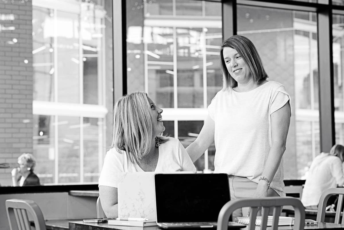 two women looking at each other with a computer in front of the woman sitting down. The photo is black and white