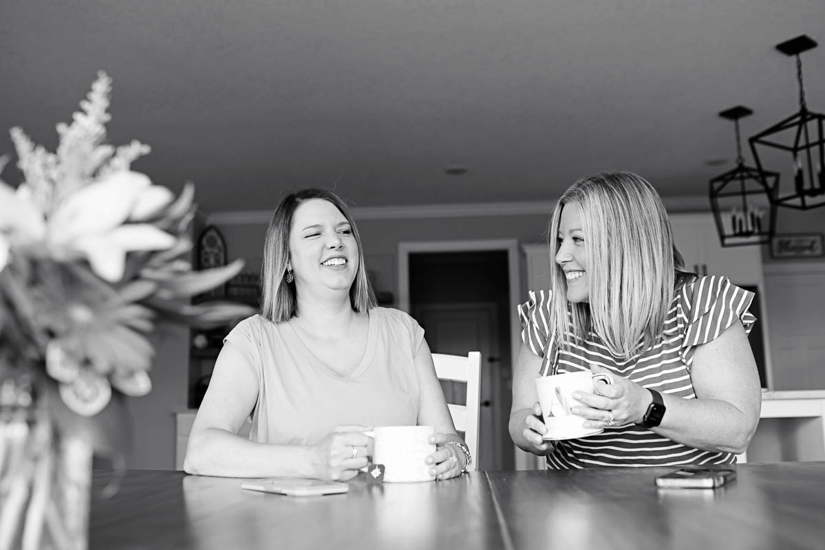 a black and white photo of two women sitting down at a table with tea and laughing
