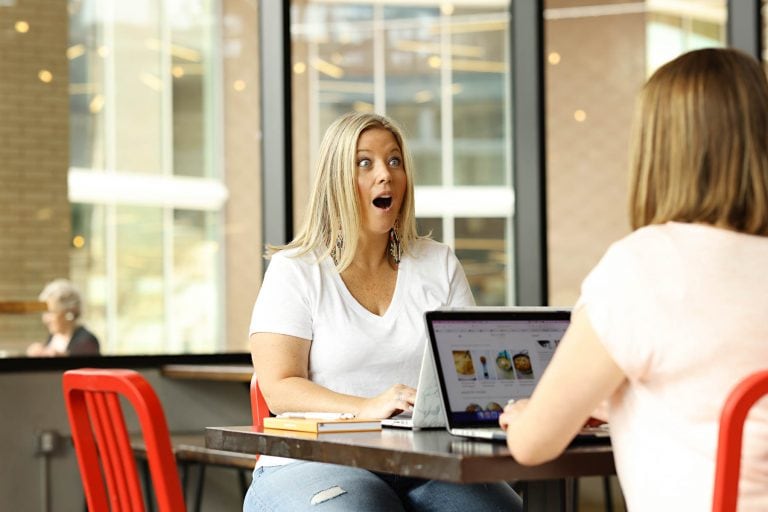 a woman with an excited face looking at a woman sitting at a computer