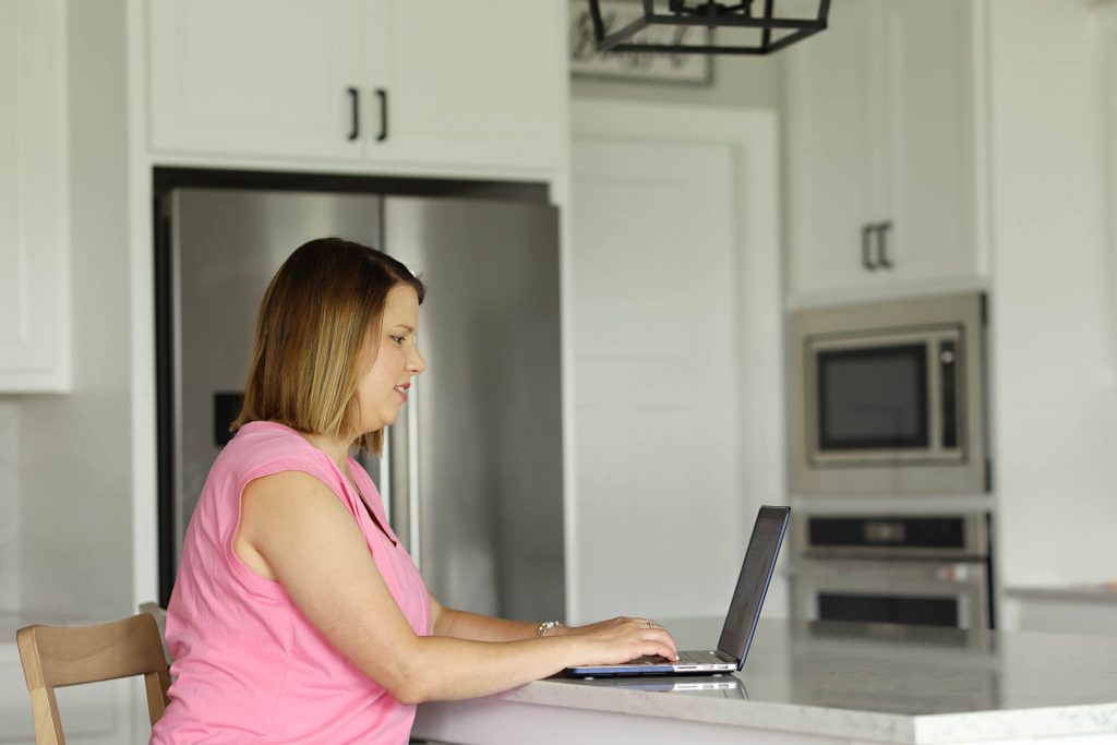 a woman in a pink shirt sitting in a kitchen typing on her computer